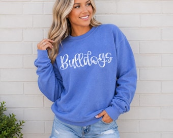 Op de afbeelding: Een blauwe sweater met ronde hals en het woord Bulldogs in een wit, verweerd script-lettertype op de borst. Het kledingstuk heeft lange mouwen, een geribbelde halslijn en een relaxte pasvorm, gecombineerd met een lichtblauwe spijkerbroekshort.