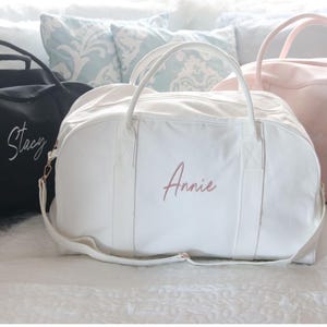 May include: Three personalized duffel bags in black, white, and pink. The white bag has the name "Annie" embroidered in a cursive font. The black bag has the name "Stacy" embroidered in a cursive font. The pink bag is plain.