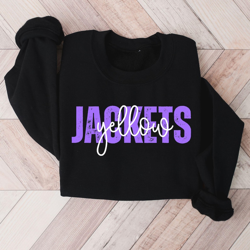 May include: Black sweatshirt with the words "JACKETS yellow" in purple and white lettering. The sweatshirt has long sleeves and a crew neck. The text has a distressed, vintage look.