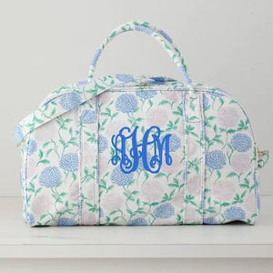 May include: A floral-patterned duffel bag with blue and pink flowers on a white background. The bag features a blue monogram in the center and matching handles. The bag is designed for travel and has a zipper closure.