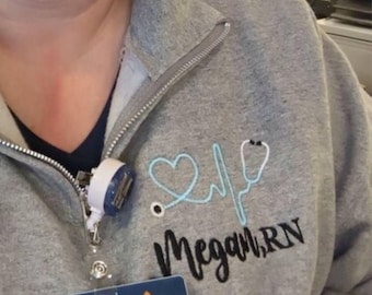 May include: Gray zippered sweatshirt with a blue and white embroidered heart and stethoscope design. The text "Megan, RN" is embroidered below the design.