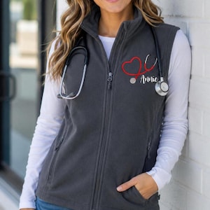 May include: A charcoal gray fleece vest featuring a full-length front zipper, a high collar, and two side pockets. Embroidered on the left chest is a red stethoscope shaped into a heart outline with the name Annie written in white script below.