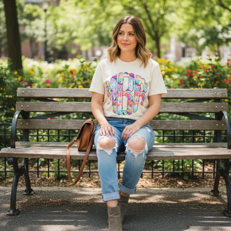 May include: A cream-colored t-shirt with a colorful monogram design. The shirt is paired with light blue ripped jeans and brown boots. A brown leather purse rests on a wooden park bench.
