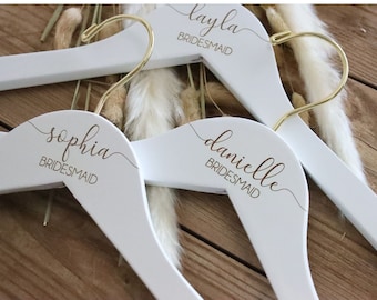 May include: Three white wooden hangers with gold hooks. Each hanger has the word "BRIDESMAID" printed on it, along with a name: "layla", "sophia", and "danielle".
