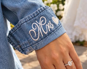 May include: A blue denim shirt cuff featuring white embroidered cursive text that reads Mrs followed by a small heart icon. The cuff is buttoned and worn on an arm adorned with a silver oval-cut diamond engagement ring on the ring finger.