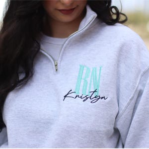 May include: A gray sweatshirt with a half-zip closure. The sweatshirt has a green embroidered design that says "RN" and "Kristyn" in a cursive font.
