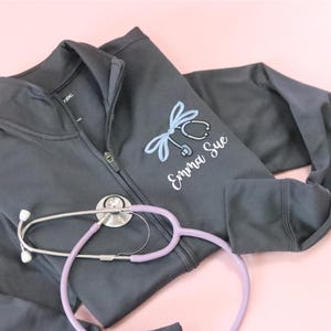 May include: A dark gray zippered jacket with a stethoscope and the name "Emma Sue" embroidered on the front. A purple stethoscope is lying on top of the jacket.