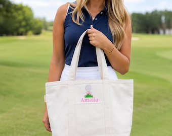 Op de afbeelding: Een crèmekleurige canvas draagtas met een geborduurd ontwerp van een golfbal op een groen grasveldje met een roze strik. De naam Amelia is in roze letters onder de golfbalafbeelding op het voorvak geborduurd.