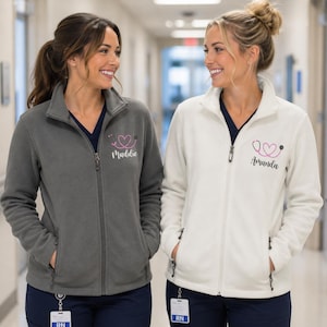 May include: Two fleece zip-up jackets, one gray and one white, with a stethoscope heart design and names printed on the chest. The jackets have a high collar, pockets, and are worn over navy blue scrubs. Both women are smiling.