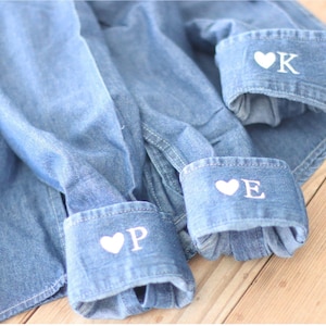 May include: Light blue denim shirt with personalized cuffs. The cuffs display white embroidered initials with a heart symbol. The shirt is laid out on a wooden surface.