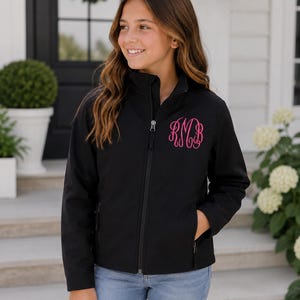 May include: A black softshell jacket featuring a high collar, a full-length front zipper, and long sleeves. The left chest area is embroidered with a bright pink monogram featuring the letters RMB in a decorative, cursive script font.