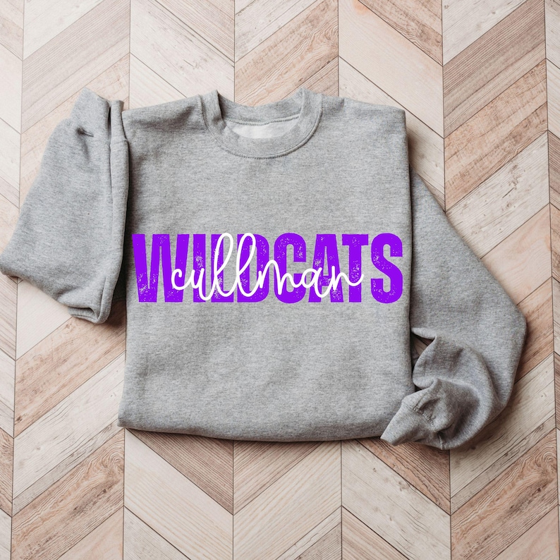 May include: A heather gray sweatshirt with the word "WILDCATS" in a distressed purple font. The word "CULLMAN" is written in a white script font over the purple text. The sweatshirt has a crew neck and long sleeves.