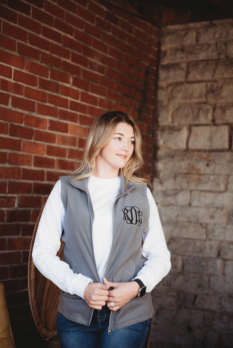 May include: A gray zip-up vest with a white long-sleeve shirt underneath. The vest has a black embroidered monogram on the front. The person is wearing blue jeans and a watch.