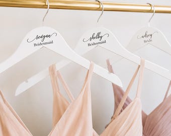 May include: Three white wooden clothes hangers with silver metal hooks hanging from a gold rod. Each hanger features black script text reading "redgan Bridesmaid", "shelby Bridesmaid", and "adrey Bride". Pale pink dresses hang from each hanger.