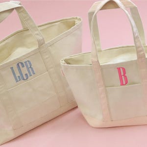 May include: Two canvas tote bags with handles. The larger bag is beige with a blue embroidered monogram "LCR". The smaller bag is beige with a pink embroidered monogram "B".