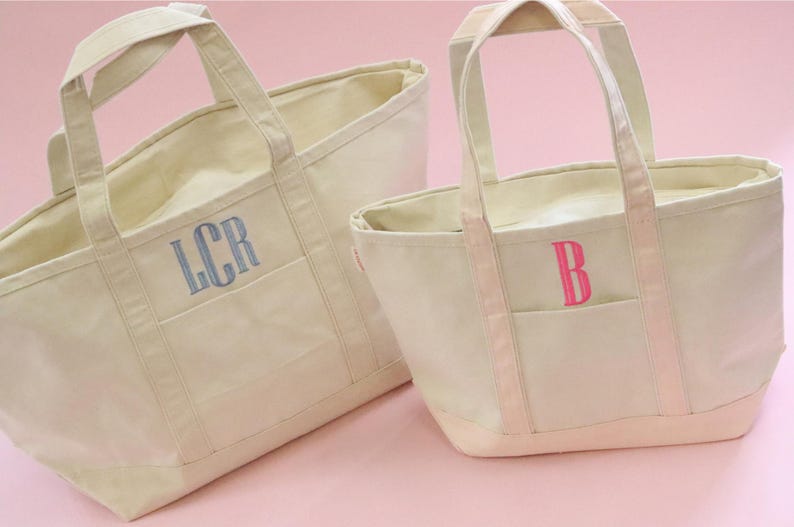 May include: Two canvas tote bags with handles. The larger bag is beige with a blue embroidered monogram "LCR". The smaller bag is beige with a pink embroidered monogram "B".
