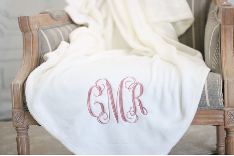 May include: White plush throw blanket with a pink embroidered monogram "GMR" on a wooden chair.