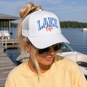 May include: White trucker hat with the words "LAKE life" in blue and red lettering. The hat has a white front panel and a white mesh back. The hat is worn by a person wearing sunglasses and a yellow shirt.
