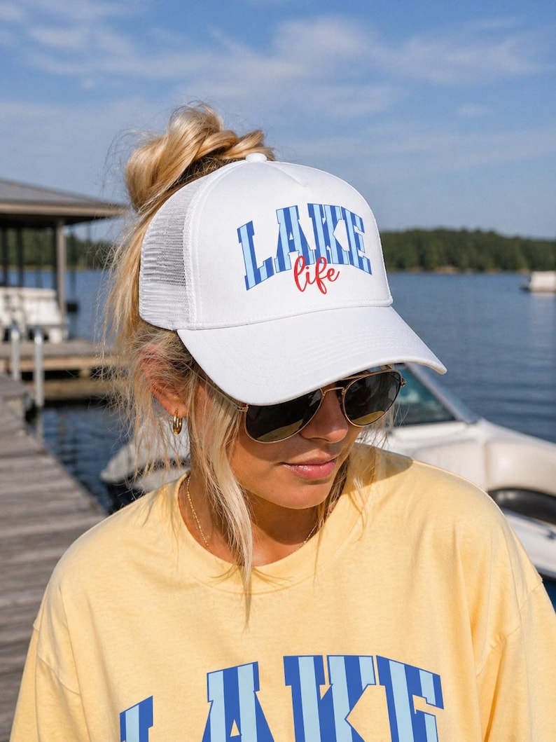 May include: White trucker hat with the words "LAKE life" in blue and red lettering. The hat has a white front panel and a white mesh back. The hat is worn by a person wearing sunglasses and a yellow shirt.