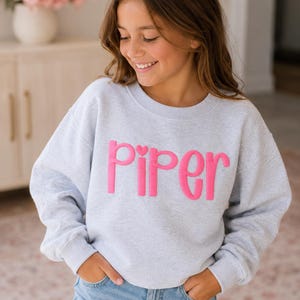 May include: A light gray crewneck sweatshirt featuring the name Piper in raised, bright pink, puffy lettering across the chest. The sweatshirt has long sleeves with ribbed cuffs and a ribbed neckline, paired with light blue denim jeans.