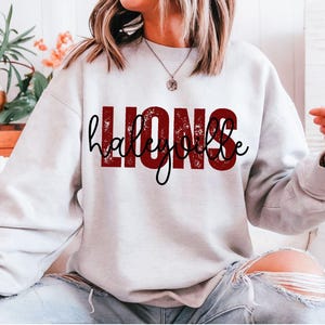 May include: A light gray sweatshirt with the word "LIONS" in a distressed red font and the name "haleybe" in a black script font. The sweatshirt is a crew neck style.
