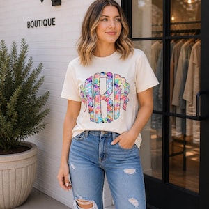 May include: Cream-colored t-shirt with a colorful tie-dye monogram design. The shirt is paired with distressed blue jeans. The word "BOUTIQUE" is visible on the building's exterior.