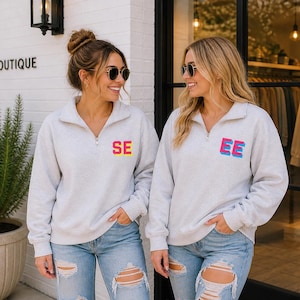 May include: Two women wearing light gray zip-up sweatshirts with the letters "SE" and "EE" in colorful block letters. They are wearing sunglasses and ripped blue jeans. The word "BOUTIQUE" is visible on the building.