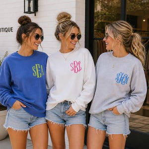 May include: Three women wearing sweatshirts and denim shorts. The sweatshirts are blue, white, and gray, each with a monogram in a contrasting color. The blue sweatshirt has "SEA" in yellow. The white sweatshirt has a pink monogram, and the gray sweatshirt has a blue monogram.