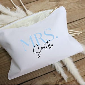 May include: White canvas cosmetic bag with a gold zipper, featuring the text "MRS. Smith" in light blue and black script. The bag is open, revealing a light interior, and rests on a wooden surface with decorative white feathers.