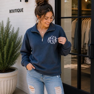 May include: A navy blue quarter-zip sweatshirt with white embroidered initials. The sweatshirt has a silver zipper and is paired with light blue distressed jeans. The word "BOUTIQUE" is visible on the building in the background.