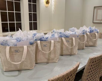 May include: A row of beige canvas tote bags with light blue monogrammed initials, filled with white tissue paper and blue floral patterned paper. The bags are arranged on a light blue table, with wicker chairs in the foreground.
