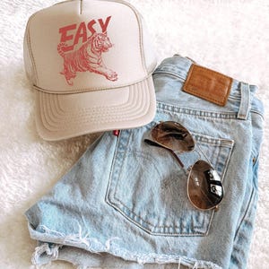 Puede incluir: Gorra de camionero beige con la palabra "EASY" y un gr&aacute;fico de tigre rojo, combinada con shorts vaqueros azul claro y gafas de sol aviador. Los shorts tienen un parche de cuero marr&oacute;n en el bolsillo trasero.