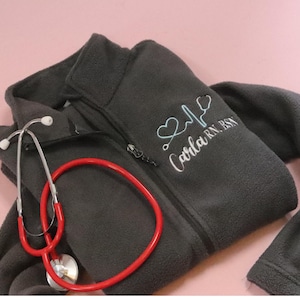 May include: Gray fleece jacket with a zipper closure and a stethoscope. The jacket has an embroidered design with a heart, a stethoscope, a heartbeat line, and the text "Carla RN BSN".