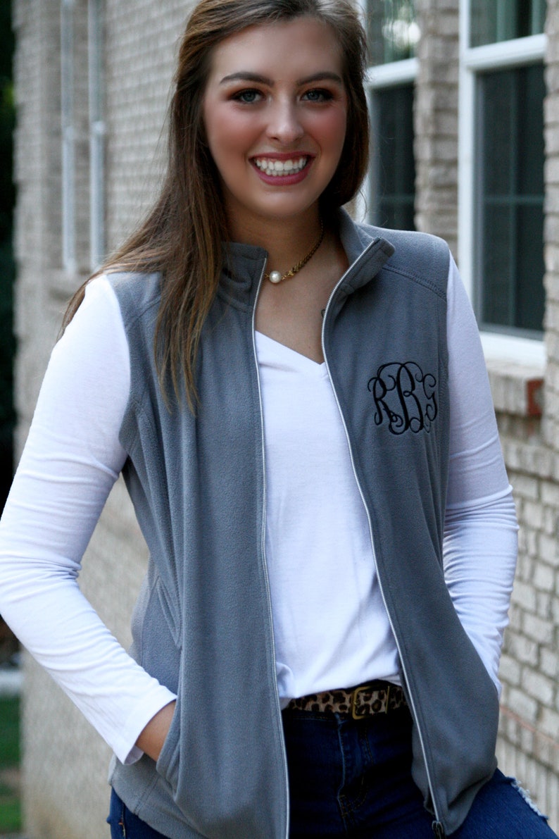 May include: A gray fleece vest with a white long-sleeve shirt underneath. The vest has a zipper closure and a monogrammed "RBG" on the front. The person is wearing a brown belt with a leopard print.
