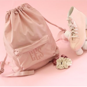 May include: A pink drawstring bag with a front pocket that says "EES" on it. The bag is next to a pair of pink roller skates with white wheels.