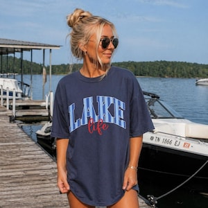 May include: Navy blue t-shirt with the word "LAKE" in blue and white block letters and the word "life" in red script. The shirt is worn on a wooden dock near a lake with boats.