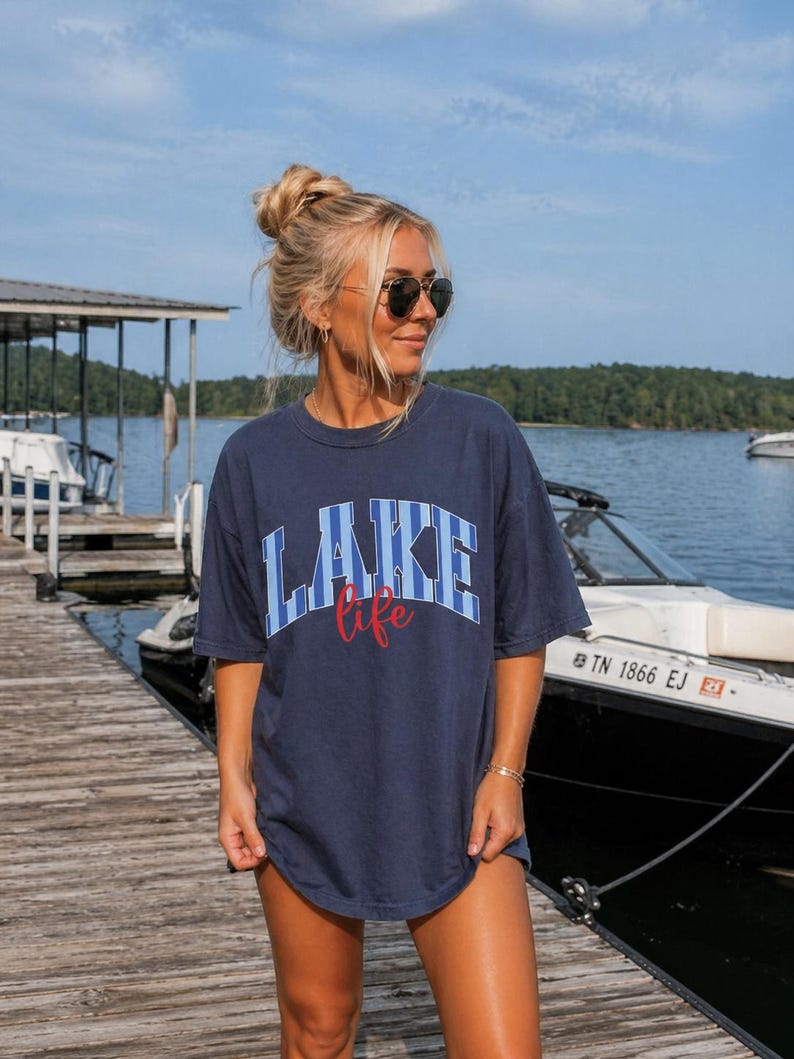 May include: Navy blue t-shirt with the word "LAKE" in blue and white block letters and the word "life" in red script. The shirt is worn on a wooden dock near a lake with boats.