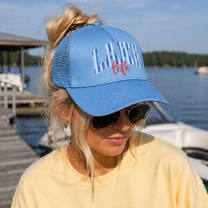 May include: A light blue trucker hat with the words "LAKE LIFE" printed on the front in white and red. The hat has a mesh back and a curved brim. The hat is worn by a person wearing sunglasses and a yellow shirt.