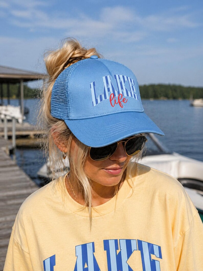 May include: A light blue trucker hat with the words "LAKE LIFE" printed on the front in white and red. The hat has a mesh back and a curved brim. The hat is worn by a person wearing sunglasses and a yellow shirt.