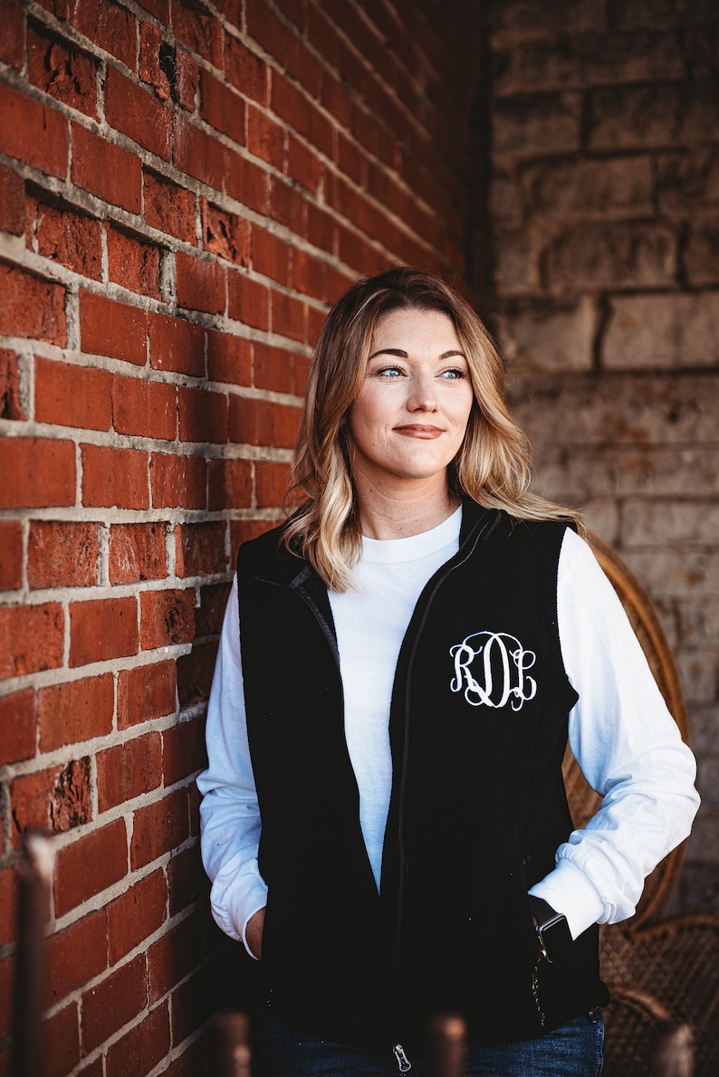 May include: A woman wearing a black fleece vest with a white long-sleeved shirt underneath. The vest has a white monogram on the front that reads "ROL".