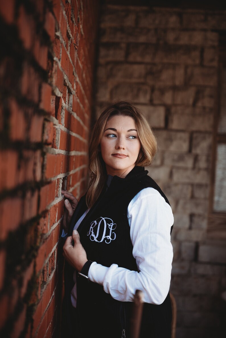 May include: A woman wearing a black fleece vest with a white monogram on the front, a white long-sleeved shirt, and dark hair. She is standing next to a brick wall.