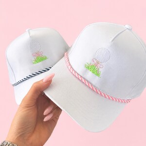 May include: Two white golf hats with embroidered golf ball and grass design. One hat has a pink and white rope brim, the other has a navy and white rope brim. The golf ball has a pink bow.