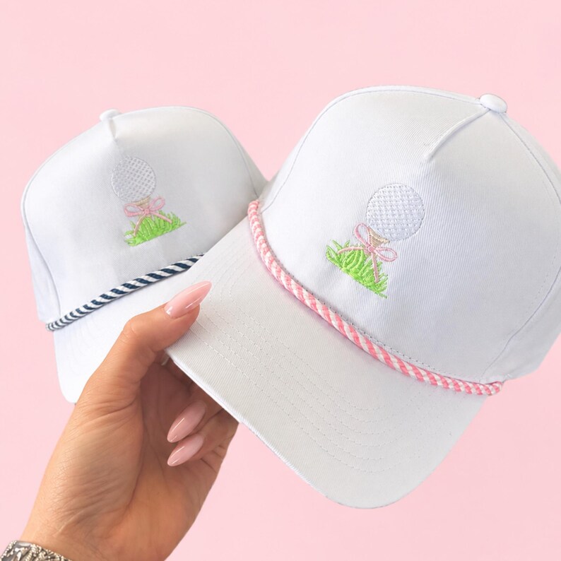 May include: Two white golf hats with embroidered golf ball and grass design. One hat has a pink and white rope brim, the other has a navy and white rope brim. The golf ball has a pink bow.