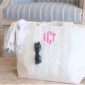 May include: A large, white canvas tote bag with a pink monogram "KCT" and a black pair of sunglasses tucked into the front pocket.