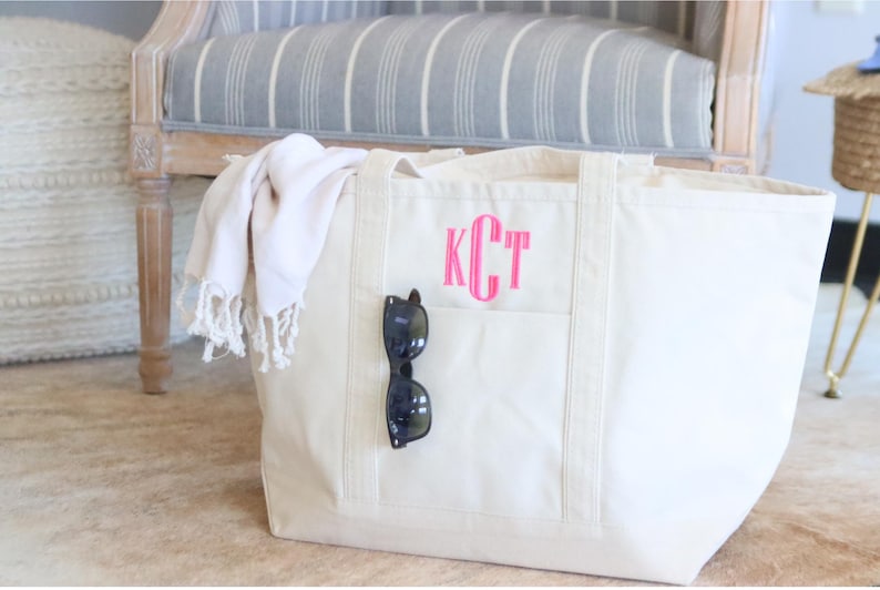 May include: A large, white canvas tote bag with a pink monogram "KCT" and a black pair of sunglasses tucked into the front pocket.