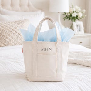 May include: A cream-colored canvas tote bag with two sturdy handles and a front pocket. The bag features the embroidered letters MHN in a gray serif font on the front. Light blue tissue paper is tucked inside the open top of the bag.