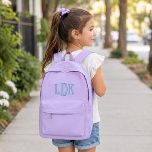 May include: A light purple backpack featuring a front zippered pocket and the embroidered monogram LDK in a teal script font. The backpack has adjustable shoulder straps and is worn on the back, showcasing a simple design with a top handle.