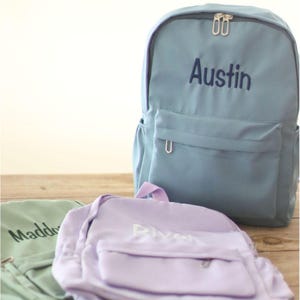 May include: Three personalized backpacks in pastel colors: light blue, lavender, and mint green. The light blue backpack has the name "Austin" embroidered in navy blue. The lavender backpack has the name "River" embroidered in white. The mint green backpack has the name "Maddox" embroidered in black.