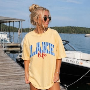 May include: Yellow t-shirt with the words "LAKE life" in blue and red lettering. The shirt is worn by a person standing on a wooden dock near a lake, with boats and a blue sky in the background.