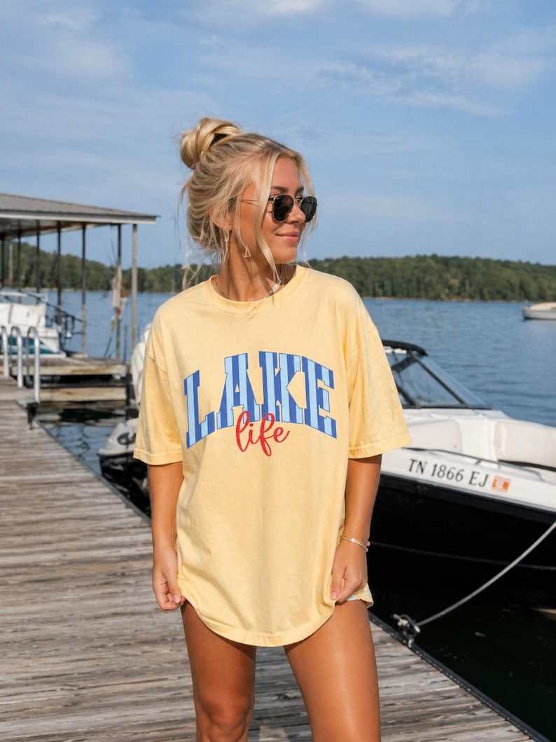 May include: Yellow t-shirt with the words "LAKE life" in blue and red lettering. The shirt is worn by a person standing on a wooden dock near a lake, with boats and a blue sky in the background.